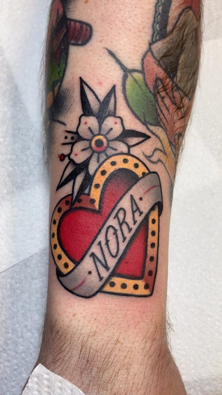 Red heart tattoo with a banner reading NORA on a person's forearm.