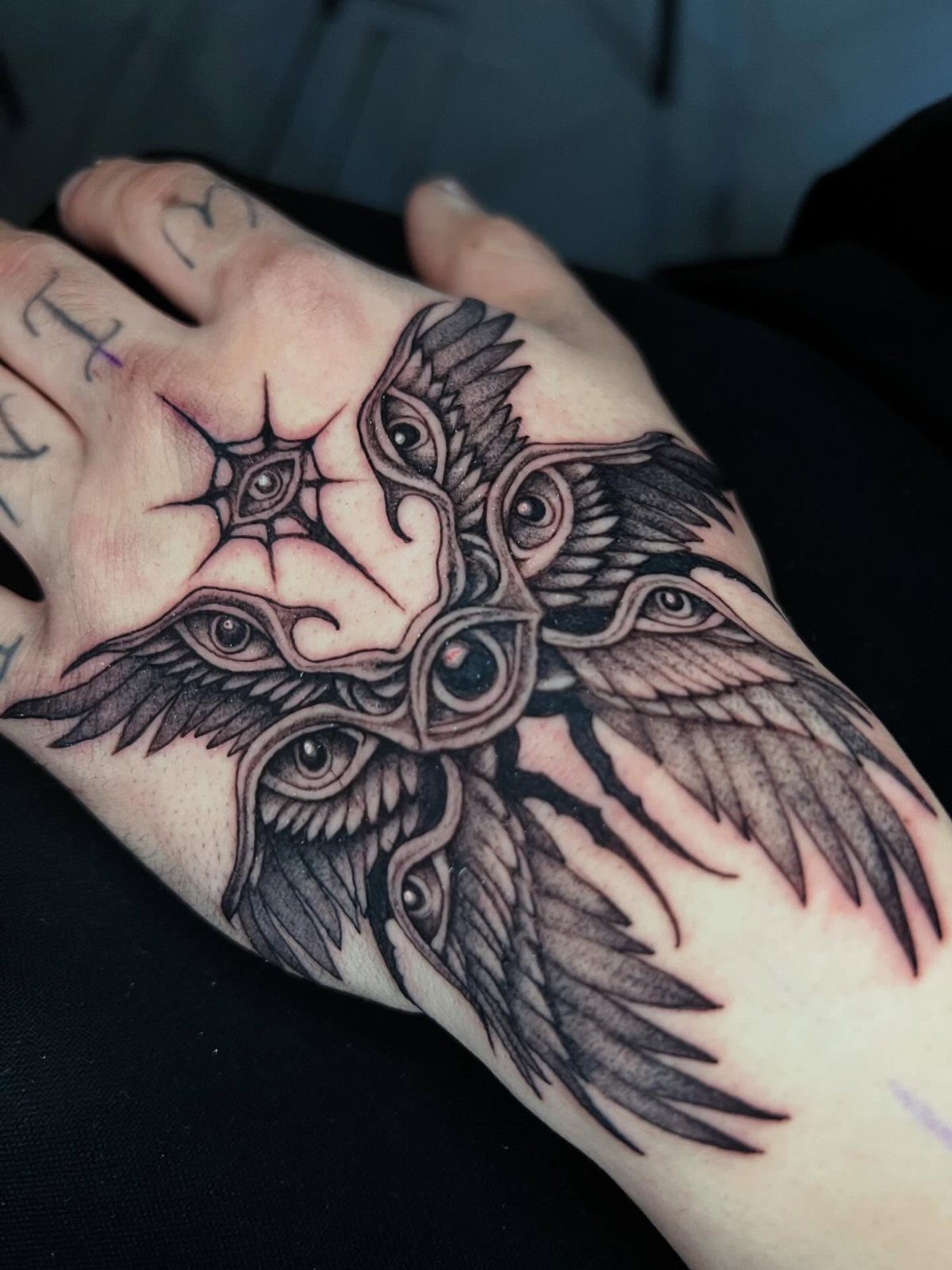Hand tattoo featuring multiple eyes surrounded by detailed black ink angel wings.
