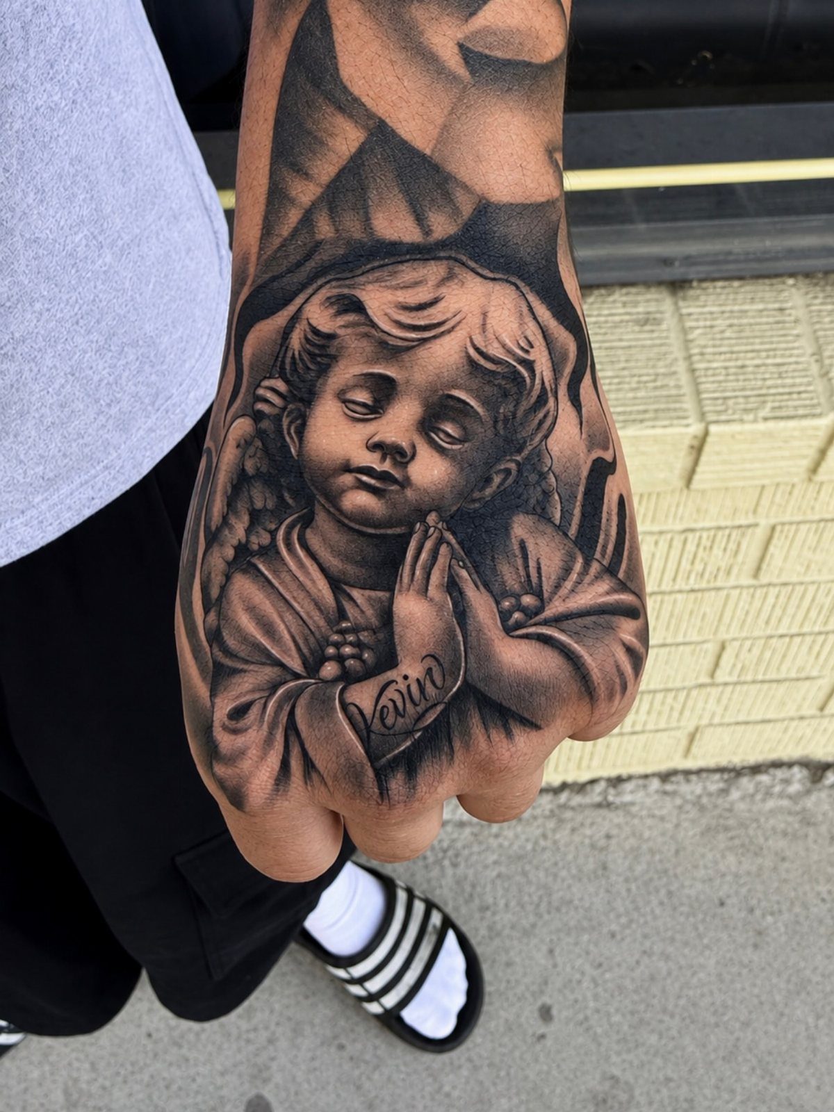 Praying cherub tattoo on a person's hand with detailed black and grey shading.