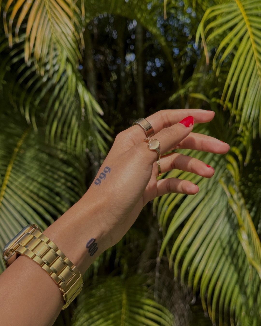 Hand with 999 angel number tattoo, gold jewelry, and green palm leaves background