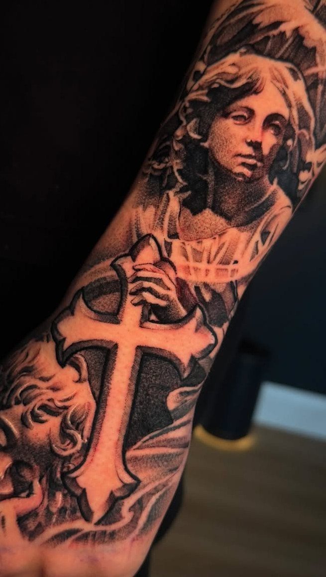 Angel and cross tattoo in black and grey ink on a forearm.