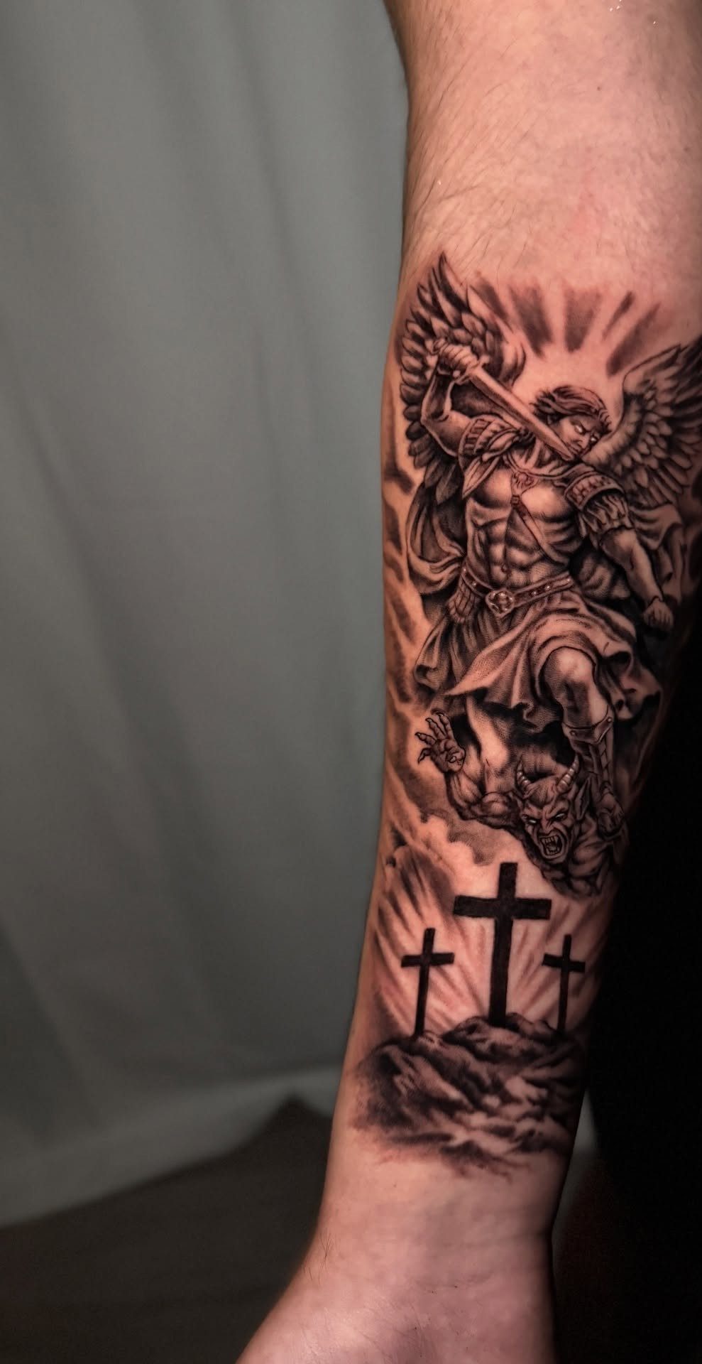 Muscular winged angel with a sword tattoo on a forearm above three crosses.