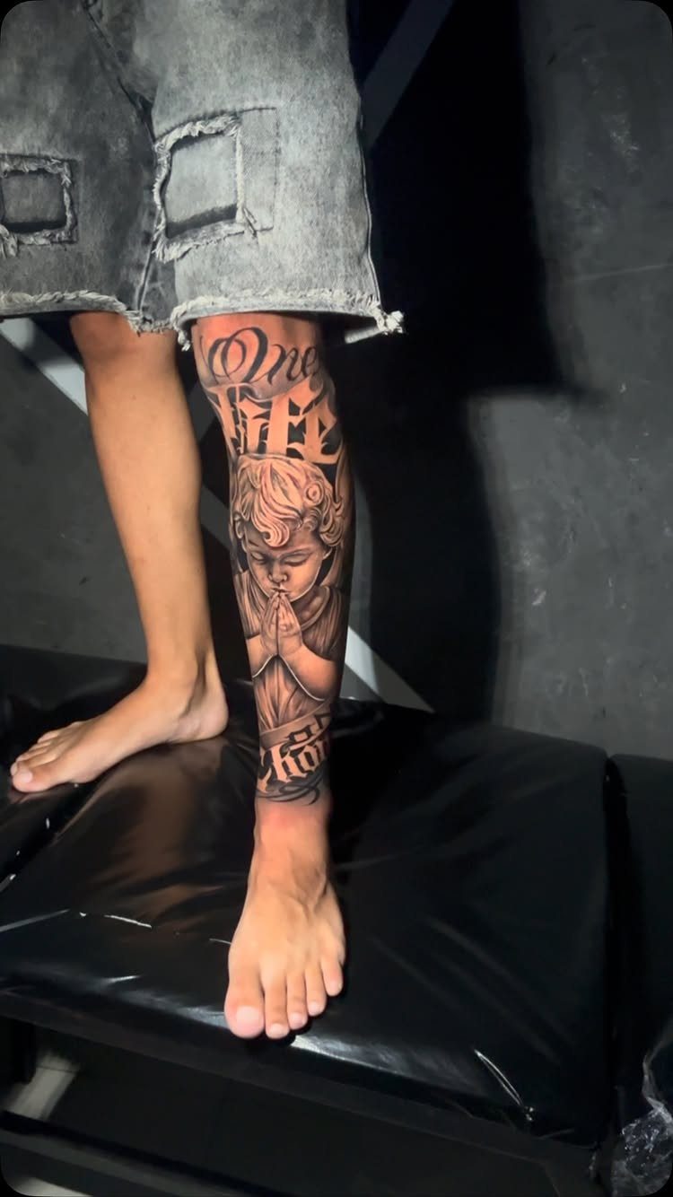 Black and grey angel tattoo on a person's lower leg with praying hands.