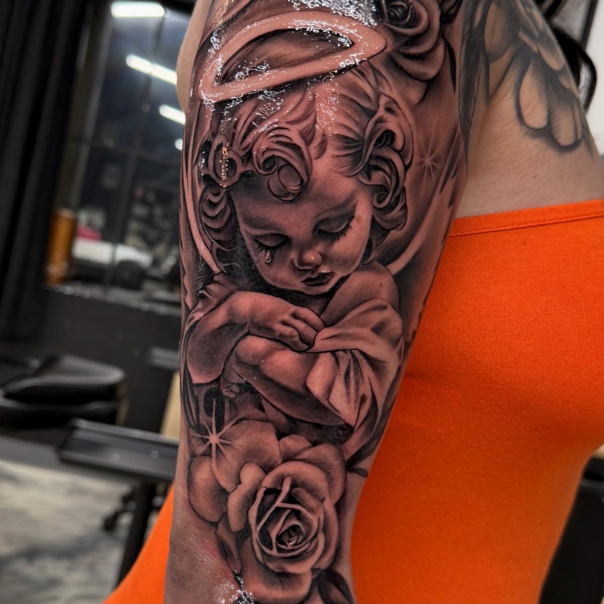 Crying cherub tattoo with a halo and a rose on an upper arm.