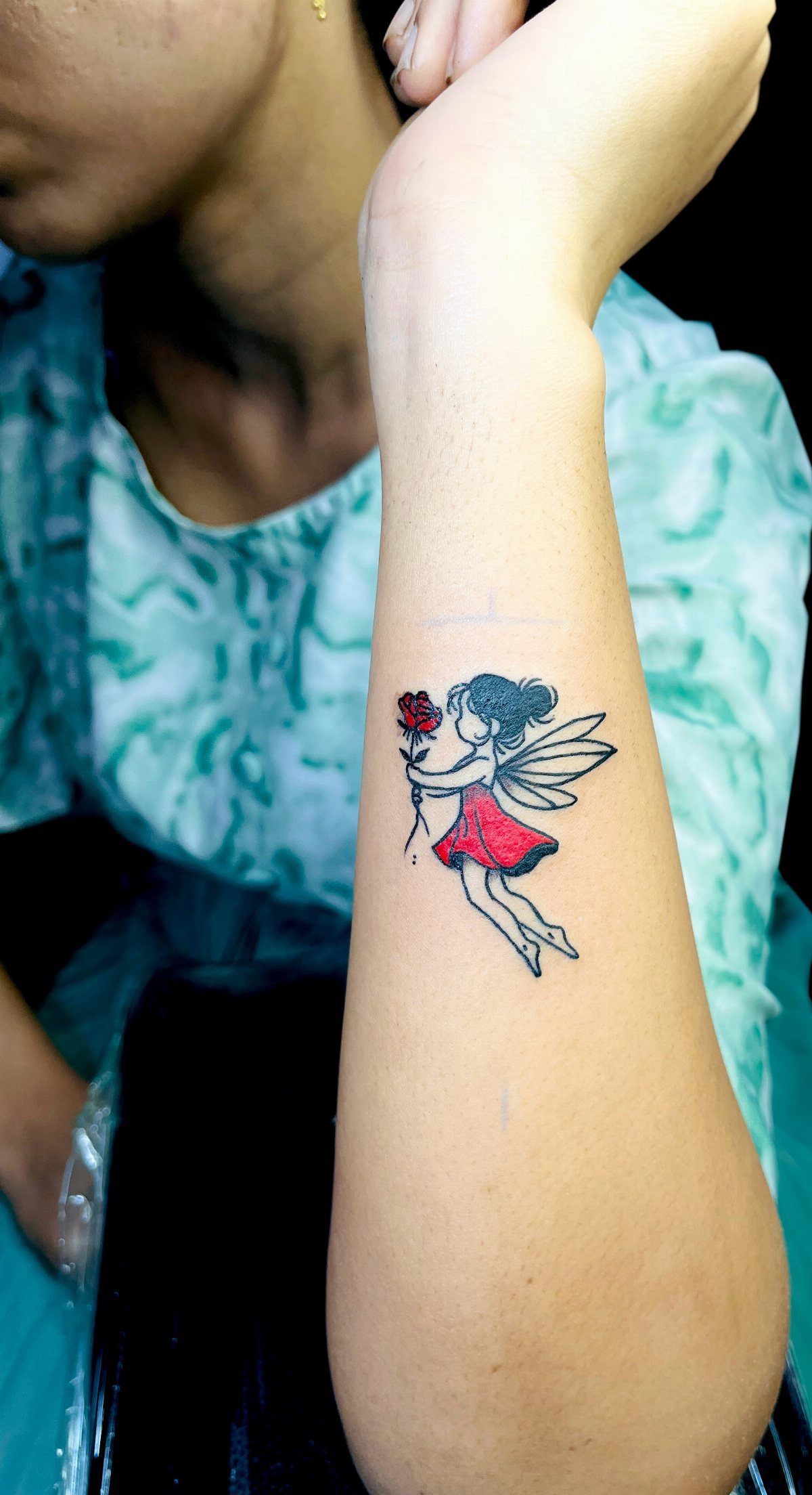 Small tattoo of a winged angel in a red dress on a forearm