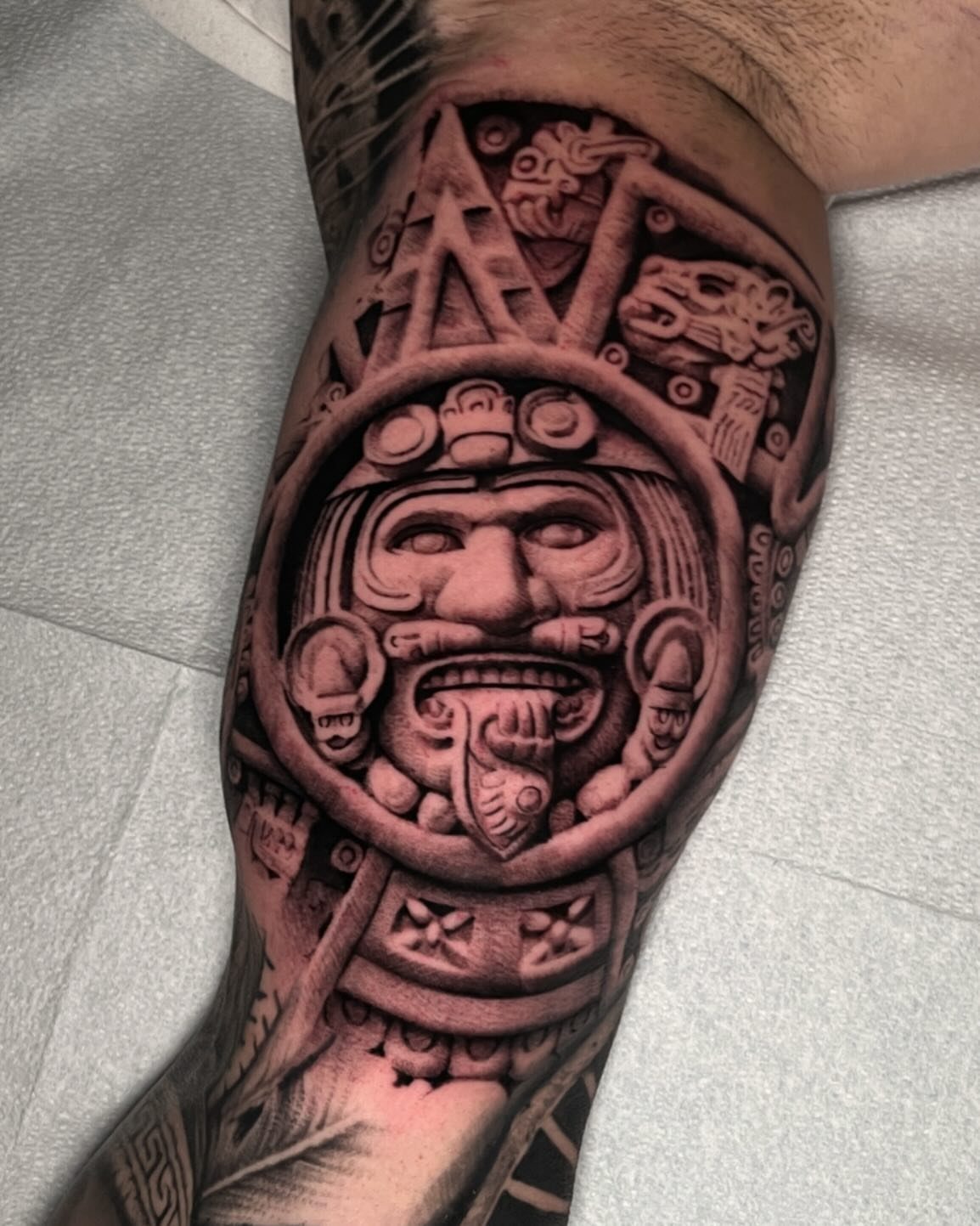 Aztec calendar stone tattoo on a forearm featuring a central face and carvings.
