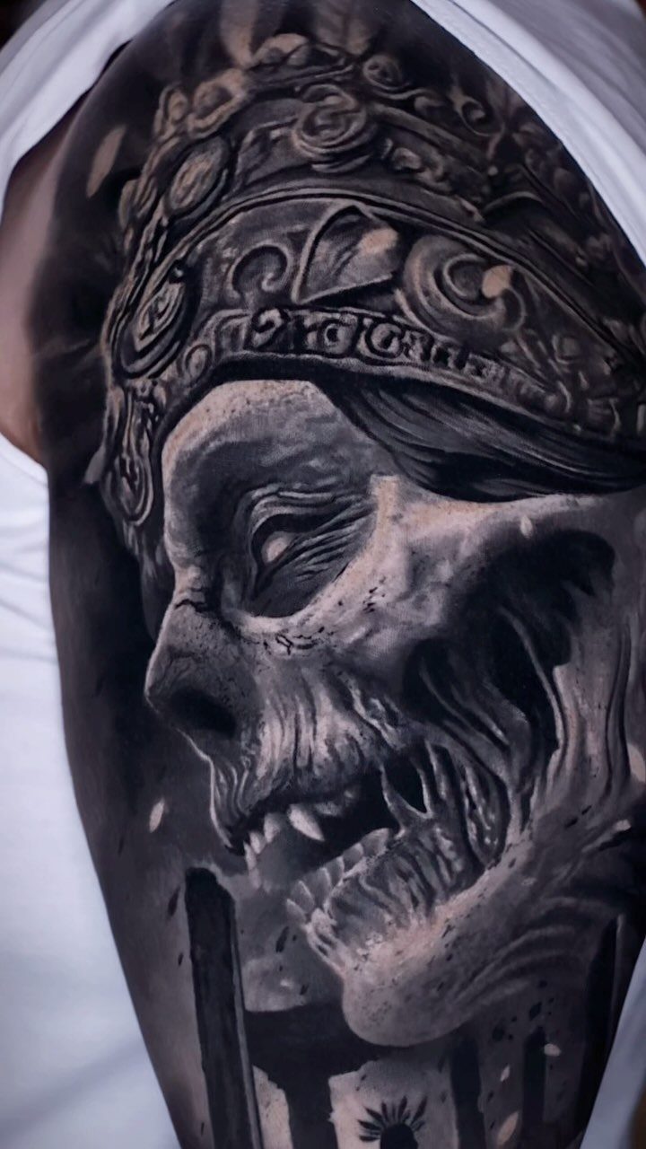Detailed black and grey tattoo of a skull wearing an ornate Aztec headdress