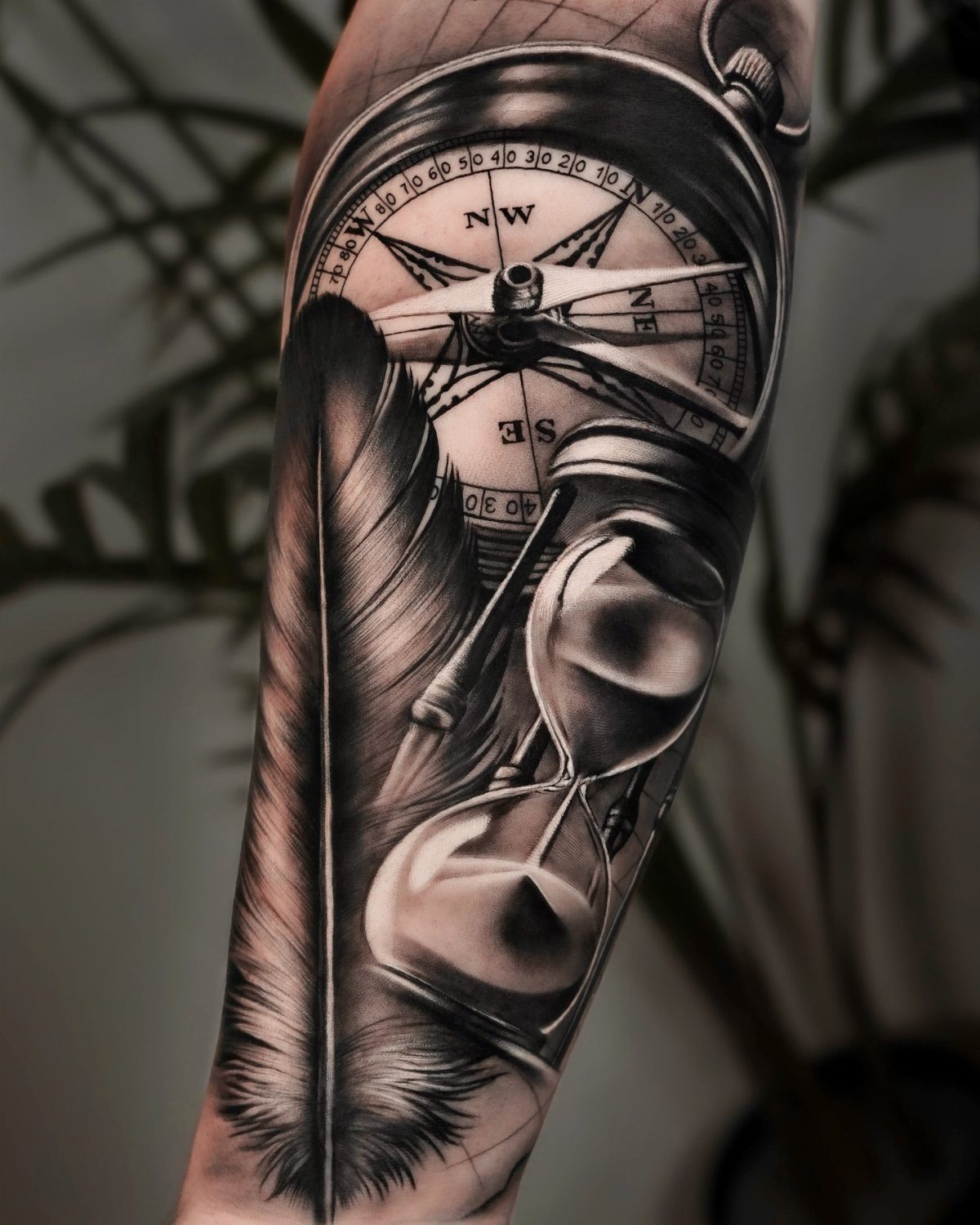 Detailed black and grey realism tattoo of a compass, hourglass, and feather on arm