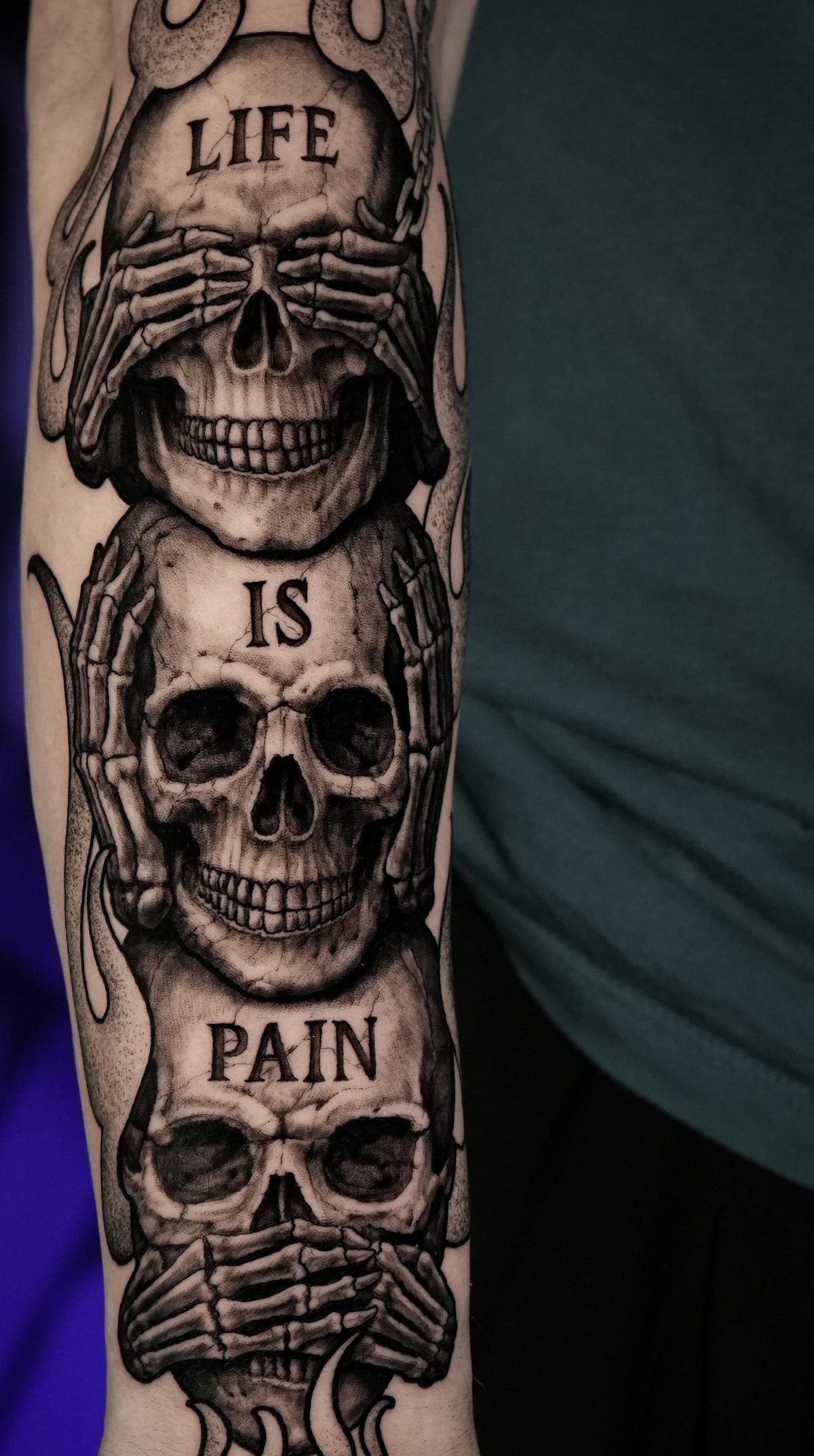Black and grey realism tattoo of three stacked skulls with skeletal hands and text.