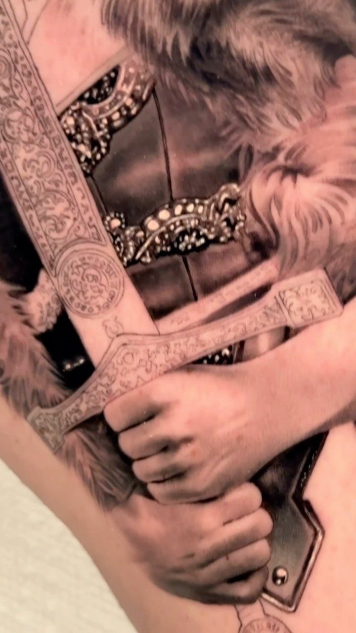 Detailed black and grey realism tattoo of hands holding an ornate sword hilt.