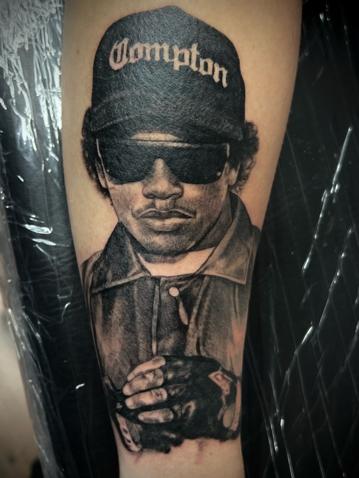 Black and grey portrait tattoo of a man wearing a Compton cap and sunglasses.