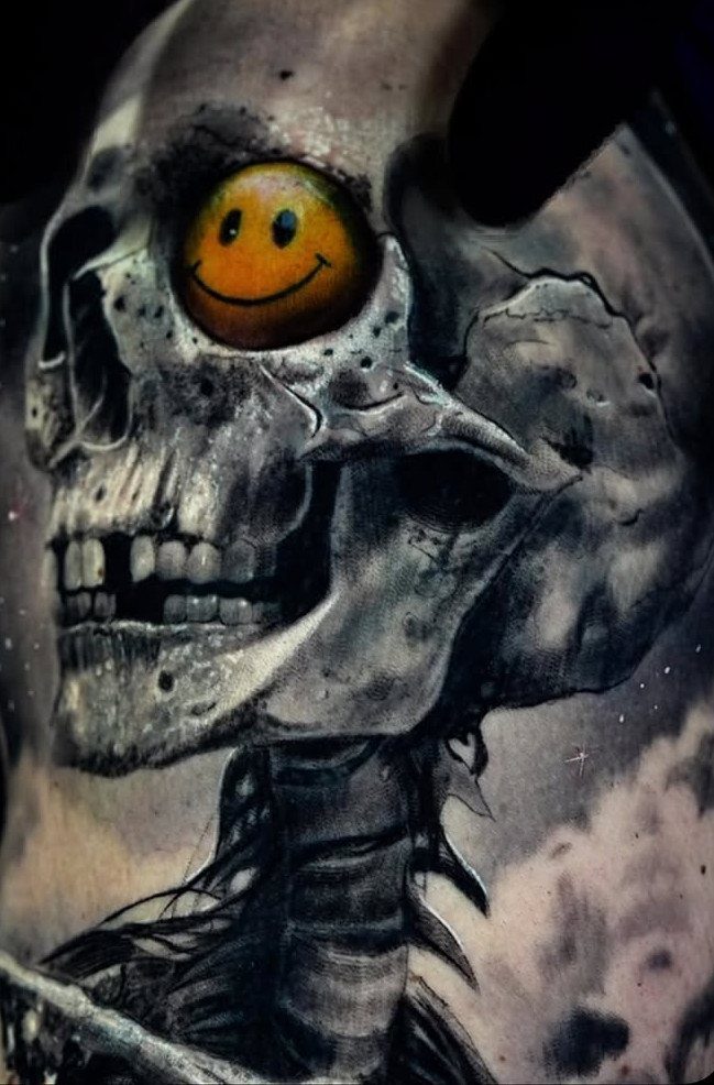 Skull tattoo in black and grey with a yellow smiley face in the eye socket