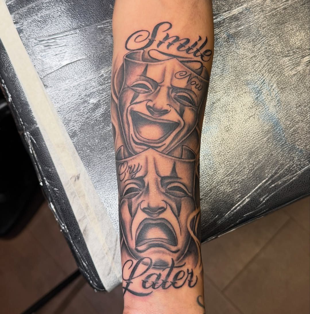 Black and grey tattoo of comedy and tragedy masks on a forearm.