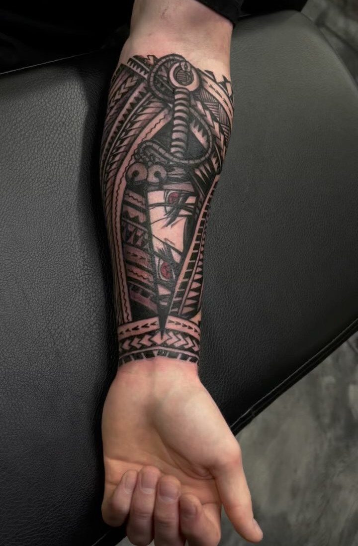 Forearm tattoo featuring bold black and grey tribal patterns and a central dagger.