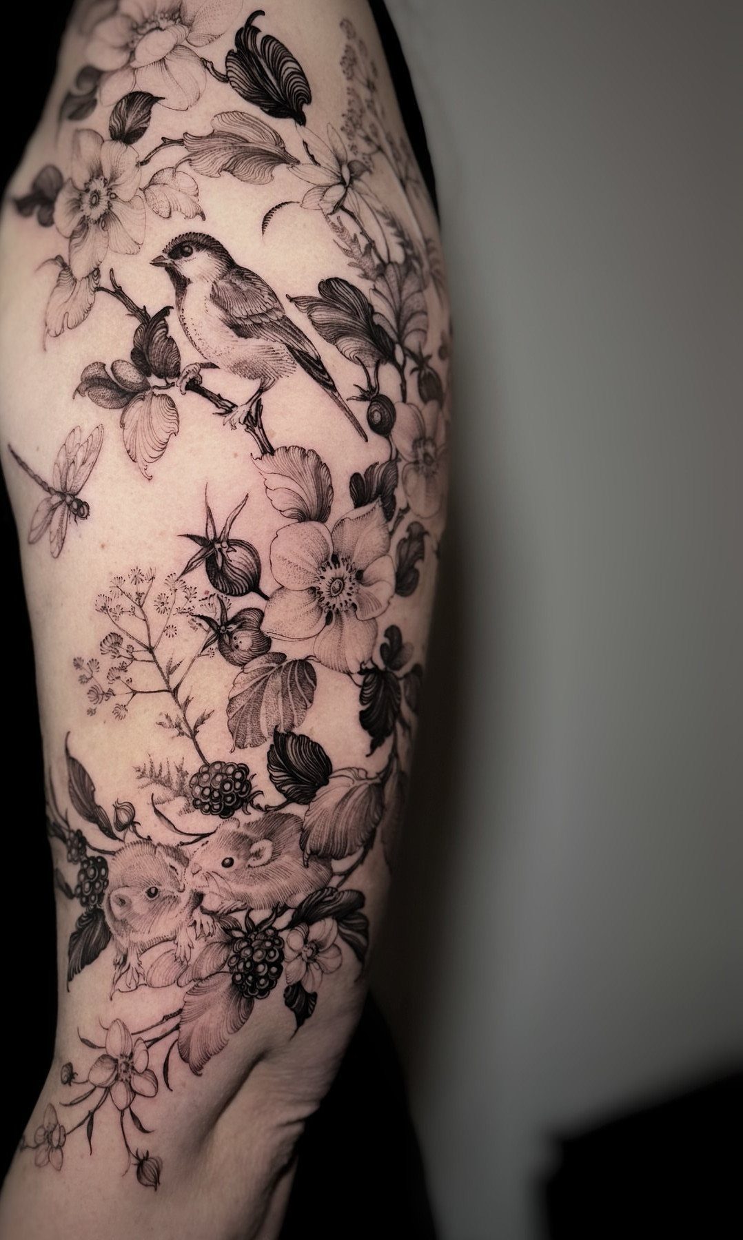 Black and grey tattoo of a bird, mice, and flowers on an arm.