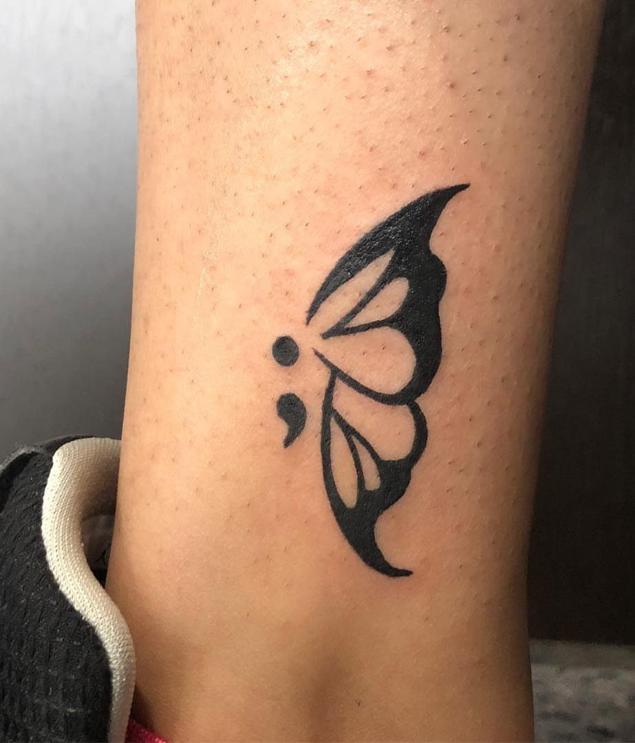 Butterfly semicolon tattoo on an ankle with a single black ink wing.