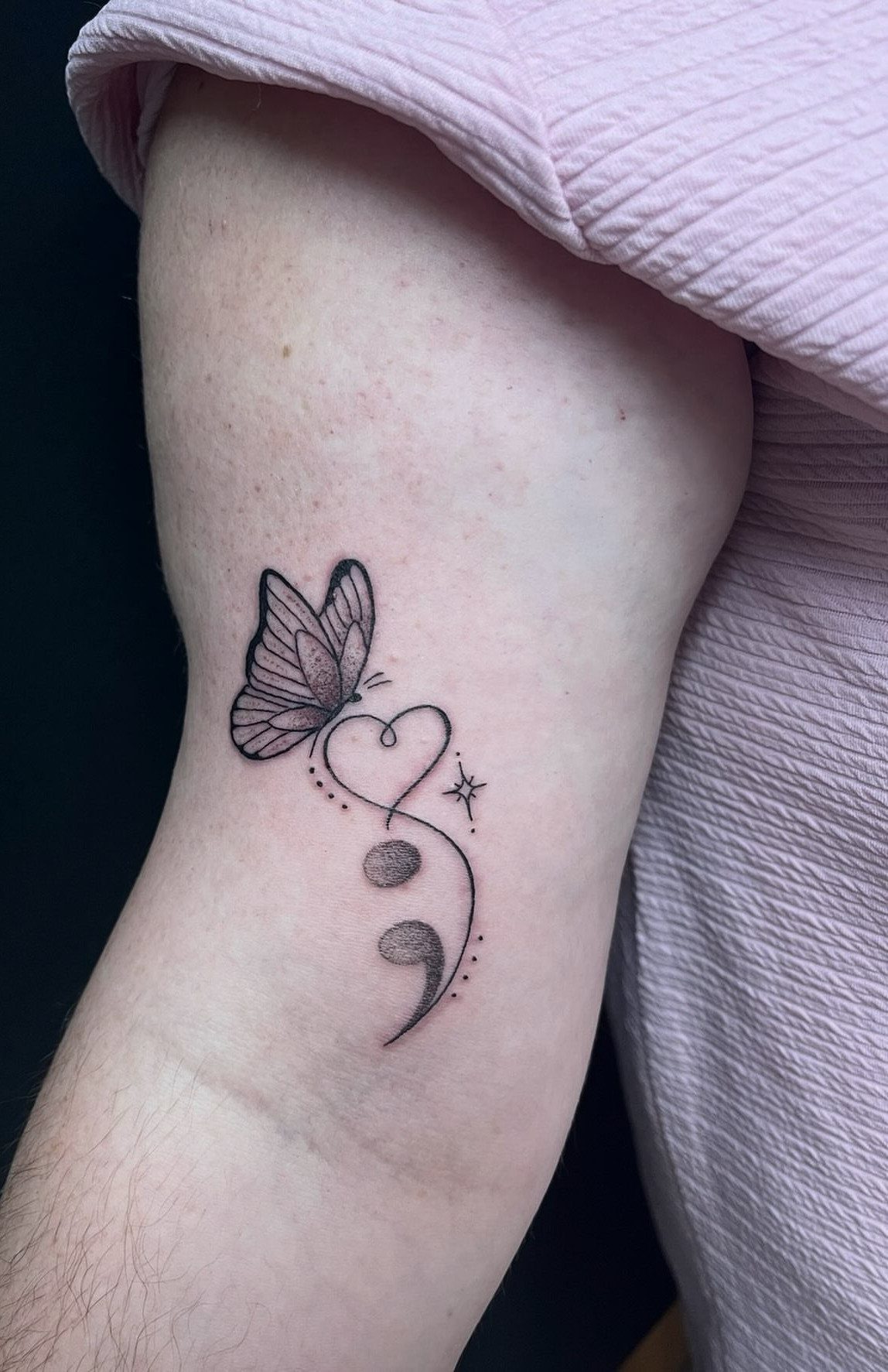 Butterfly semicolon tattoo with a heart on a person's upper arm.