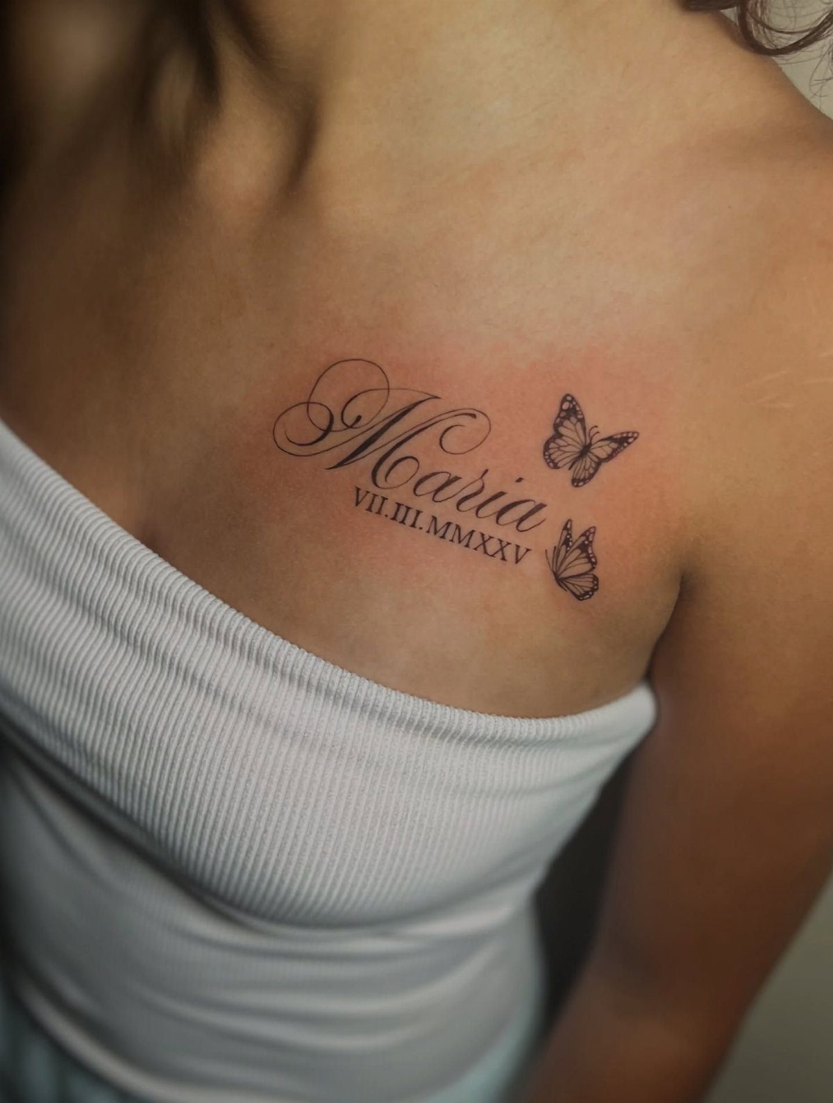Tattoo on collarbone with cursive name Maria, two butterflies, and Roman numeral date.