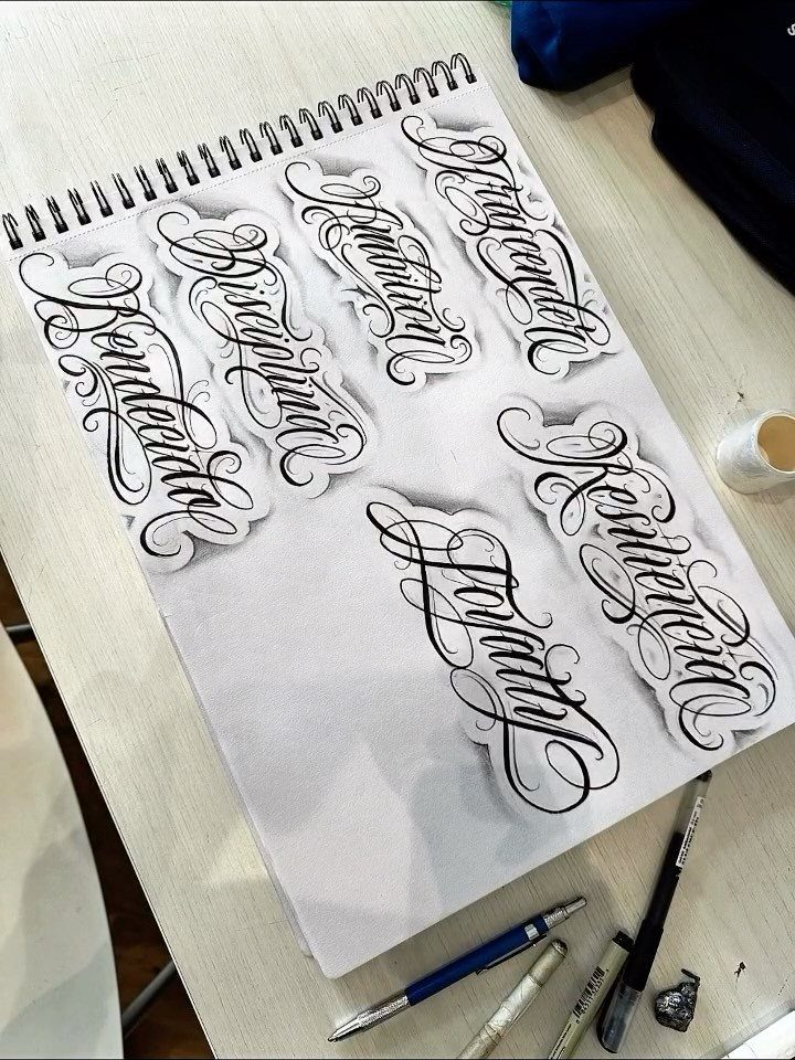 Ornate black ink calligraphy designs on a white spiral notebook with drawing pens.
