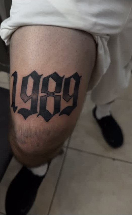 Old English style tattoo of the numbers 1989 on a person's thigh.