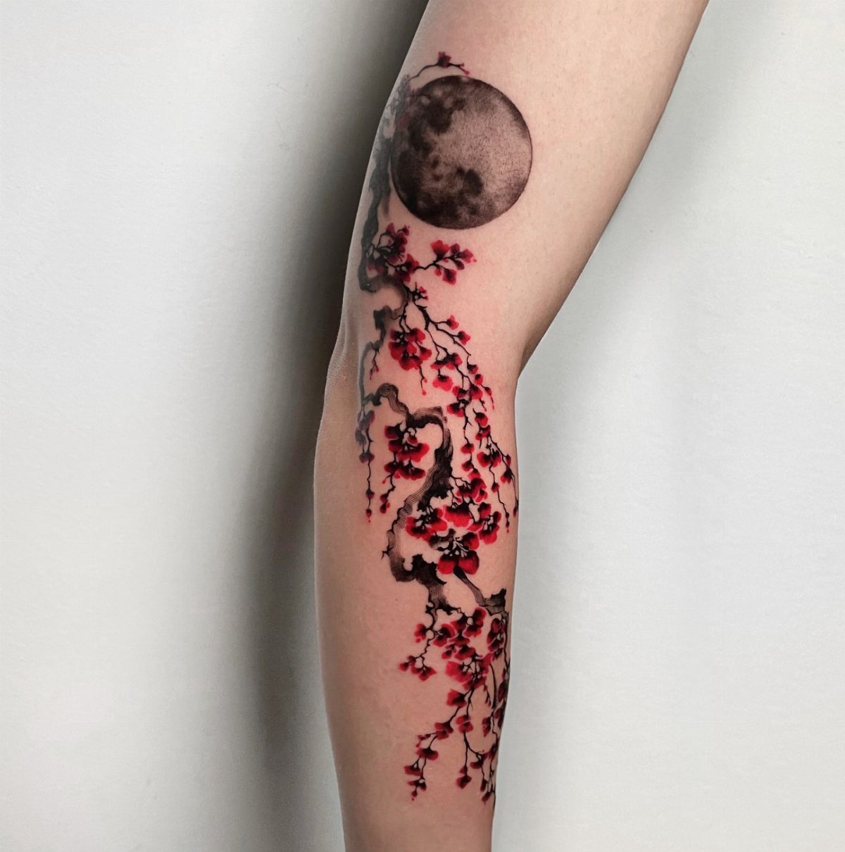 Forearm tattoo of a detailed grey moon and red cherry blossom branches.