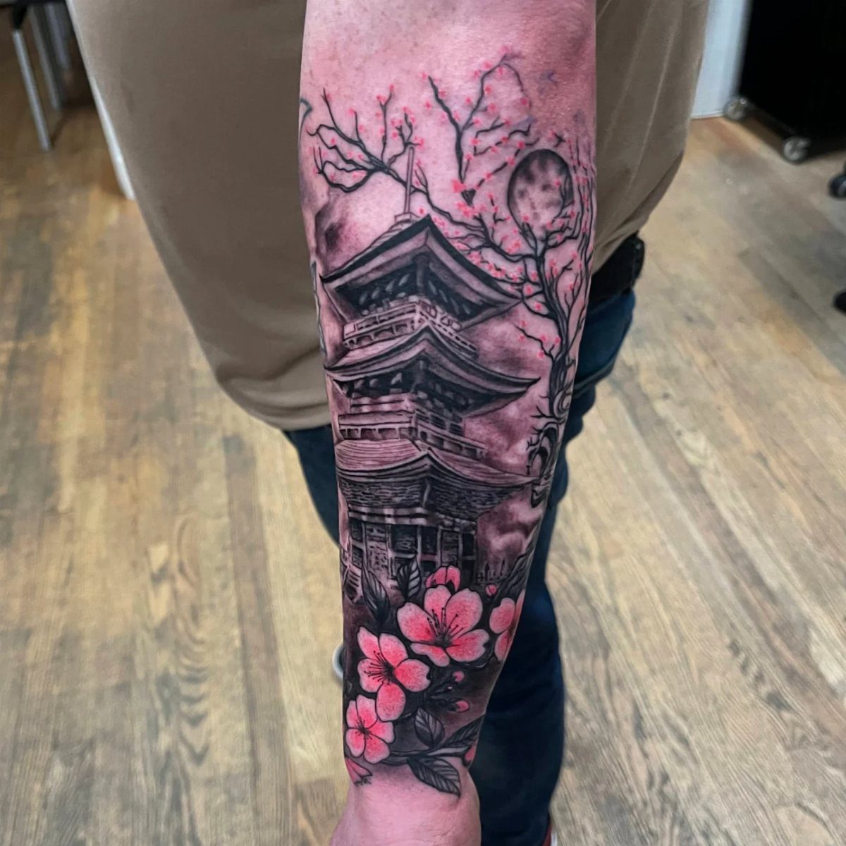 Forearm tattoo of a Japanese pagoda with pink cherry blossoms and a moon.
