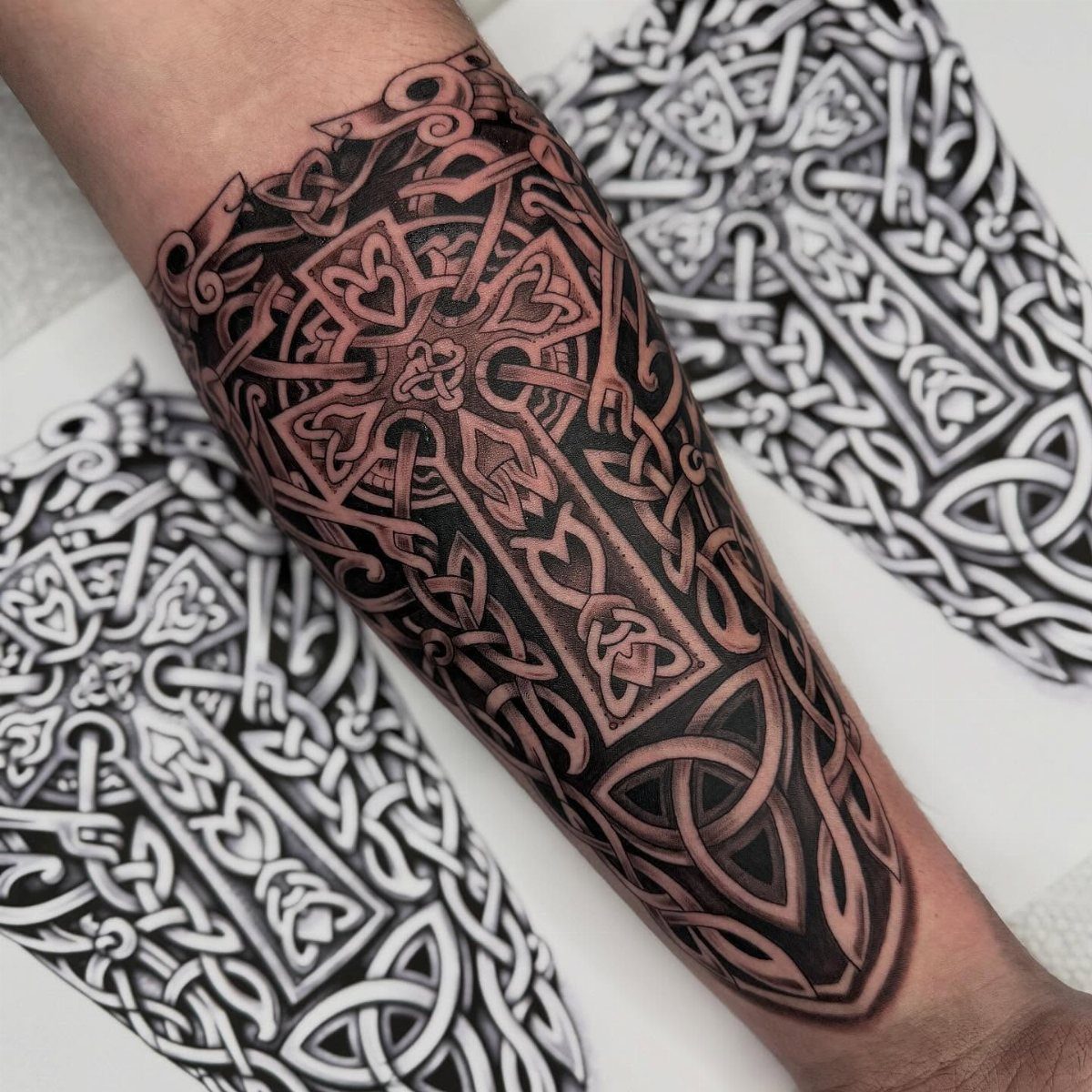 Celtic cross tattoo with intricate knotwork on a person's forearm over design sketches