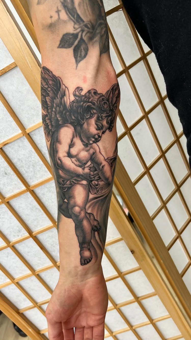 Cherub angel tattoo on a forearm with detailed wings and grey shading.