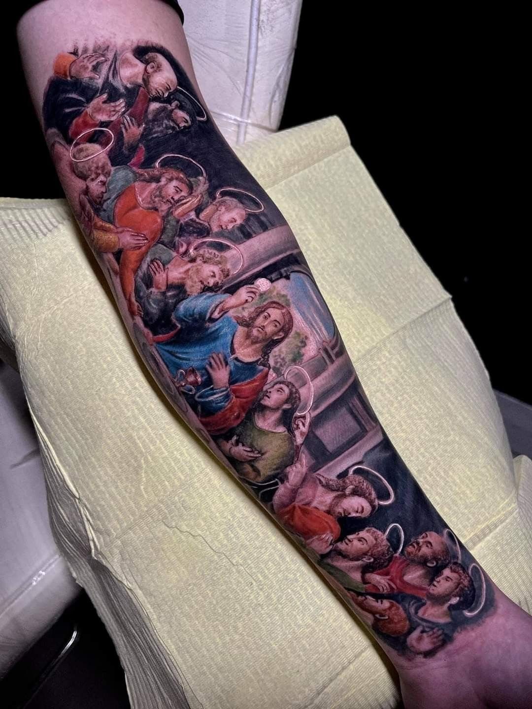 Last Supper realism tattoo on a forearm featuring Jesus and disciples with halos.