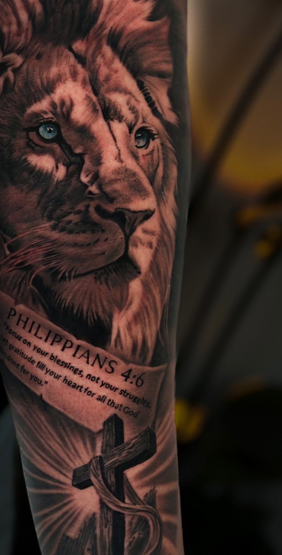 Lion tattoo with blue eyes, biblical scripture banner, and a cross on an arm.