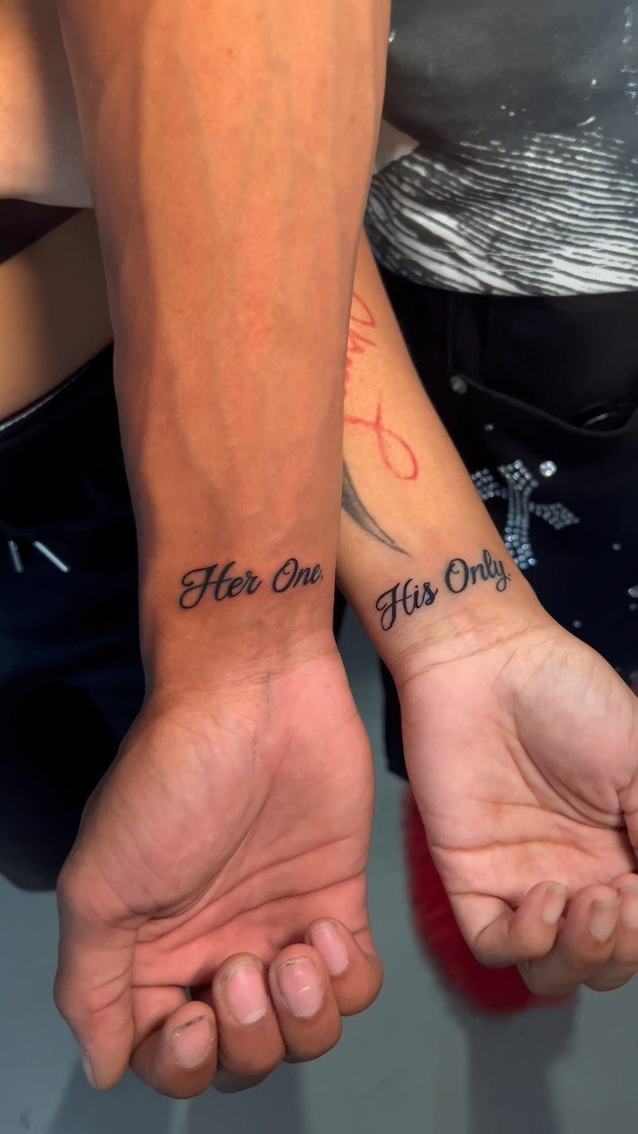 Two wrists with matching script tattoos reading Her One and His Only.