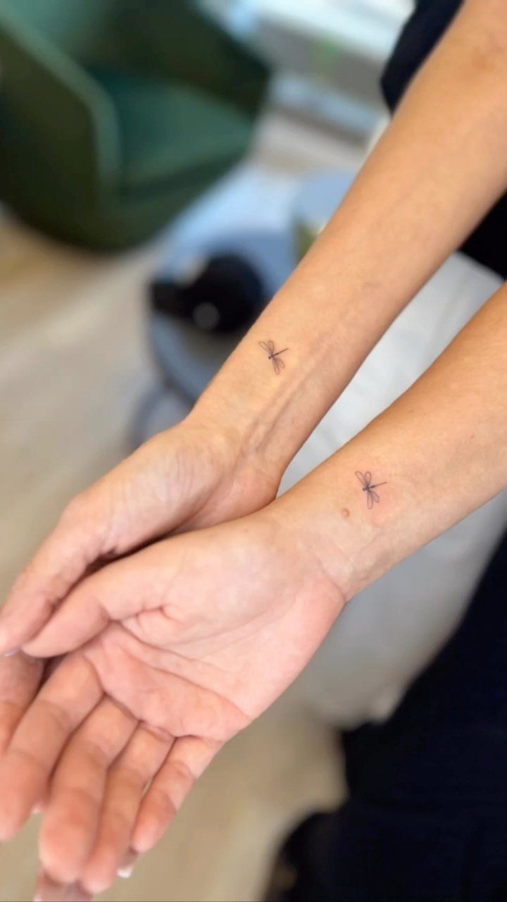 Two inner wrists each featuring a small, matching black ink dragonfly tattoo.