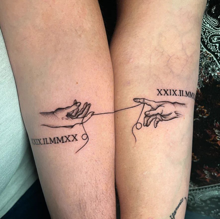 Two forearms with matching tattoos of reaching hands and Roman numeral dates.