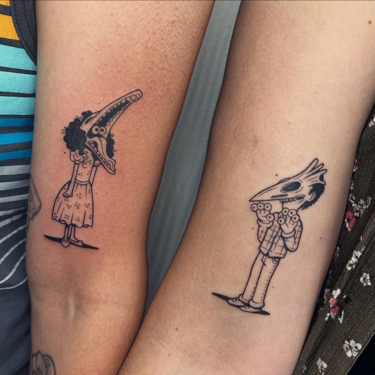 Two forearms with matching black ink tattoos of whimsical, stylized couple characters.