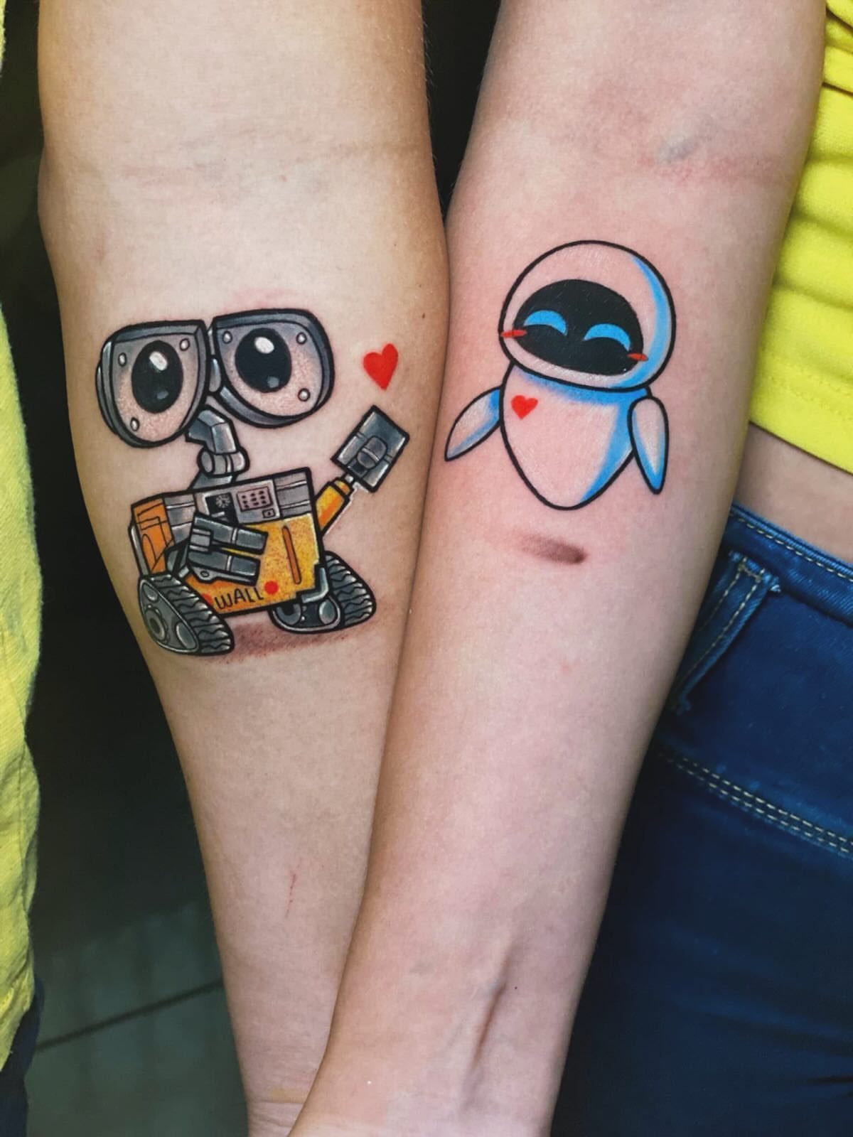 Wall-E and Eve tattoos on two forearms with a small red heart between them