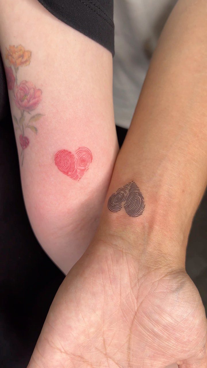Two wrists with matching heart tattoos created from fingerprint patterns in red and black.
