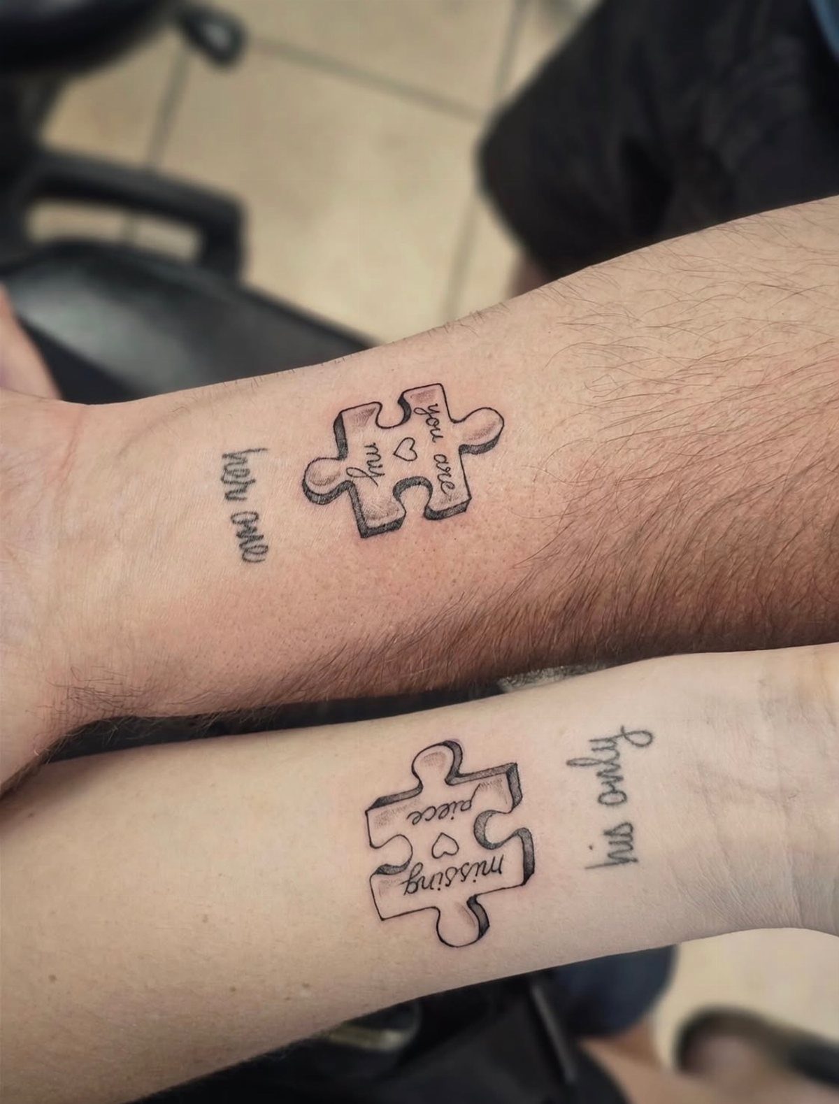 Two forearms with matching puzzle piece tattoos and romantic text in black ink.