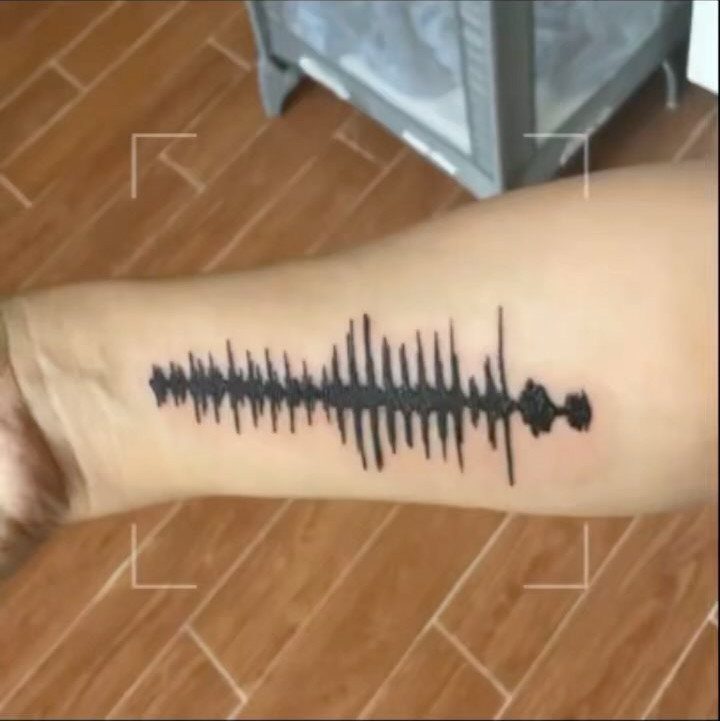 Black ink heartbeat line tattoo on a person's forearm over wooden flooring
