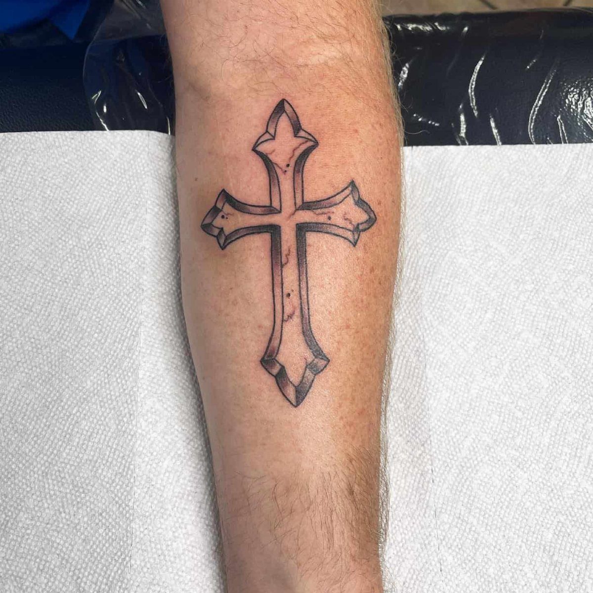 Stylized black outlined cross tattoo on a person's forearm over white paper.