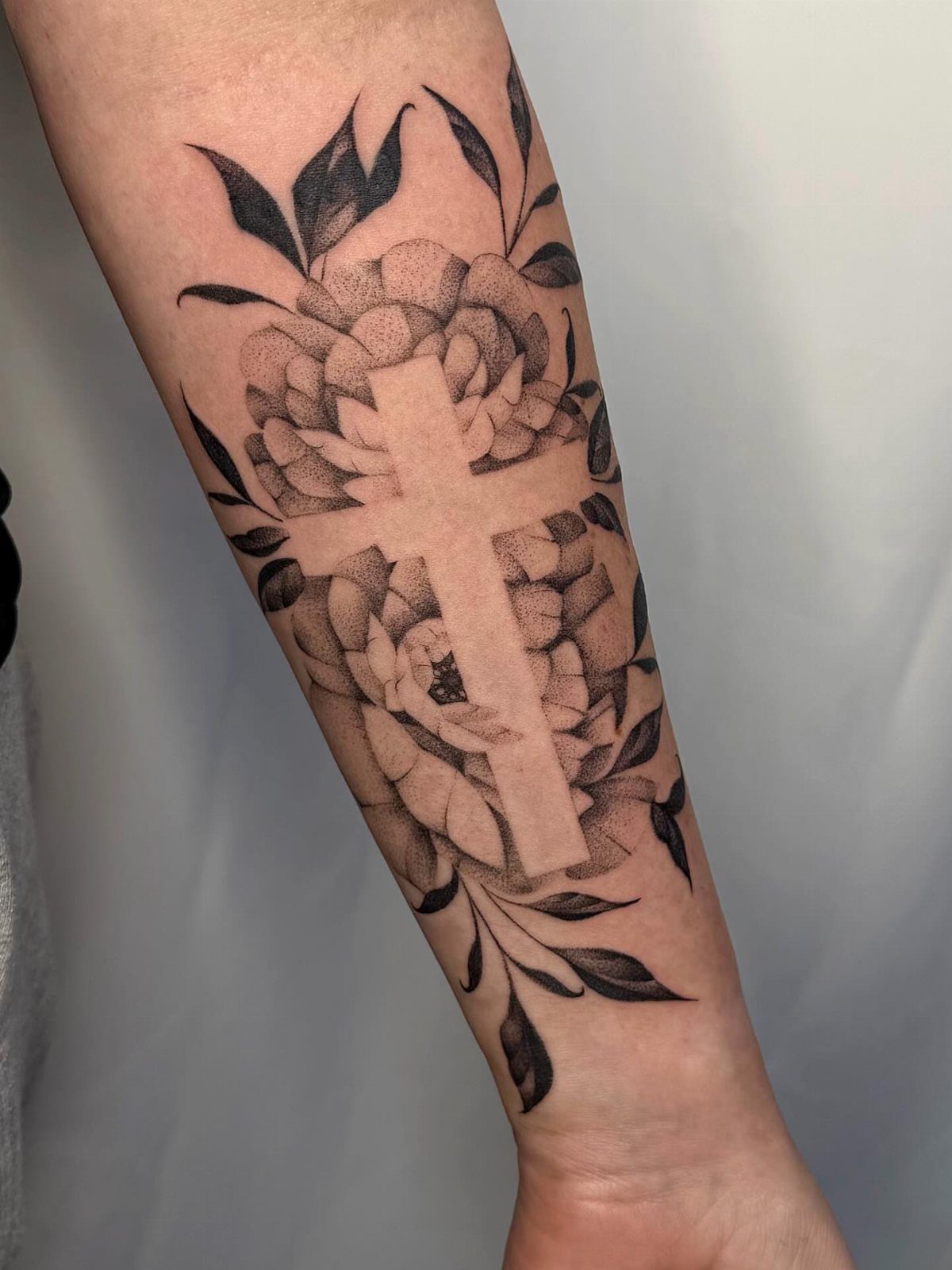 Forearm tattoo of a negative space cross surrounded by shaded flowers and leaves.