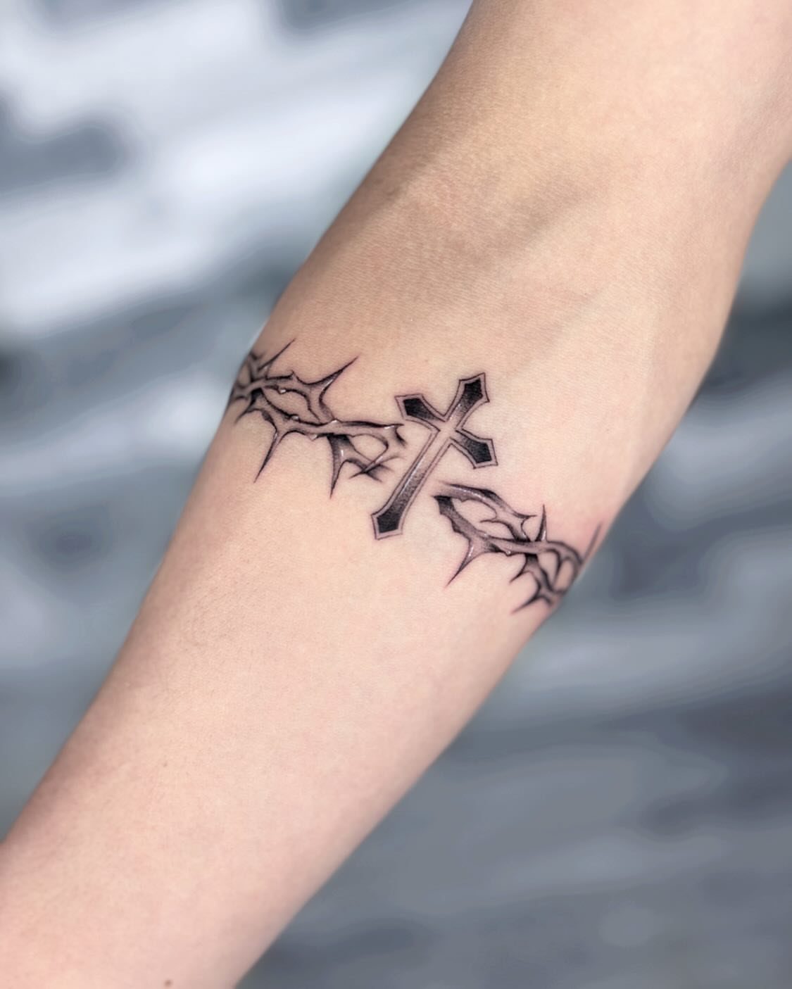 Forearm tattoo of a black cross pendant hanging from a crown of thorns band