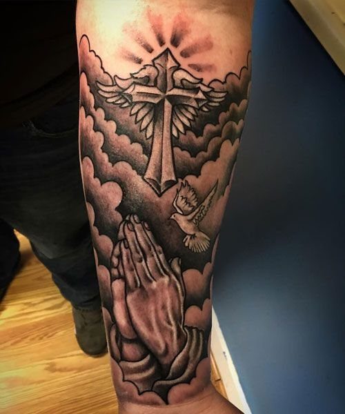 Grey-shaded forearm tattoo of a winged cross, praying hands, and a dove.