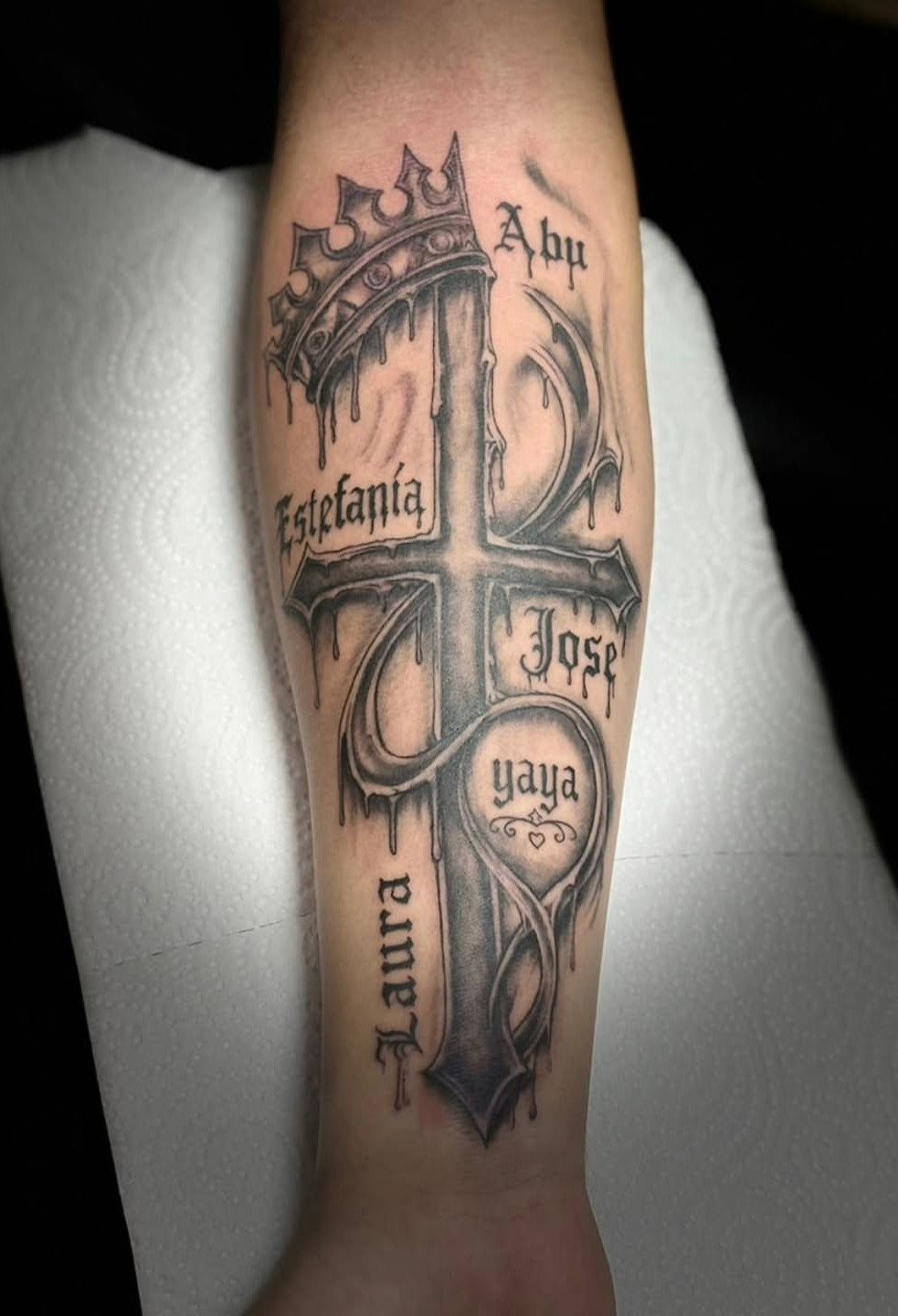 Ornate cross tattoo on a forearm featuring a crown and several written names.