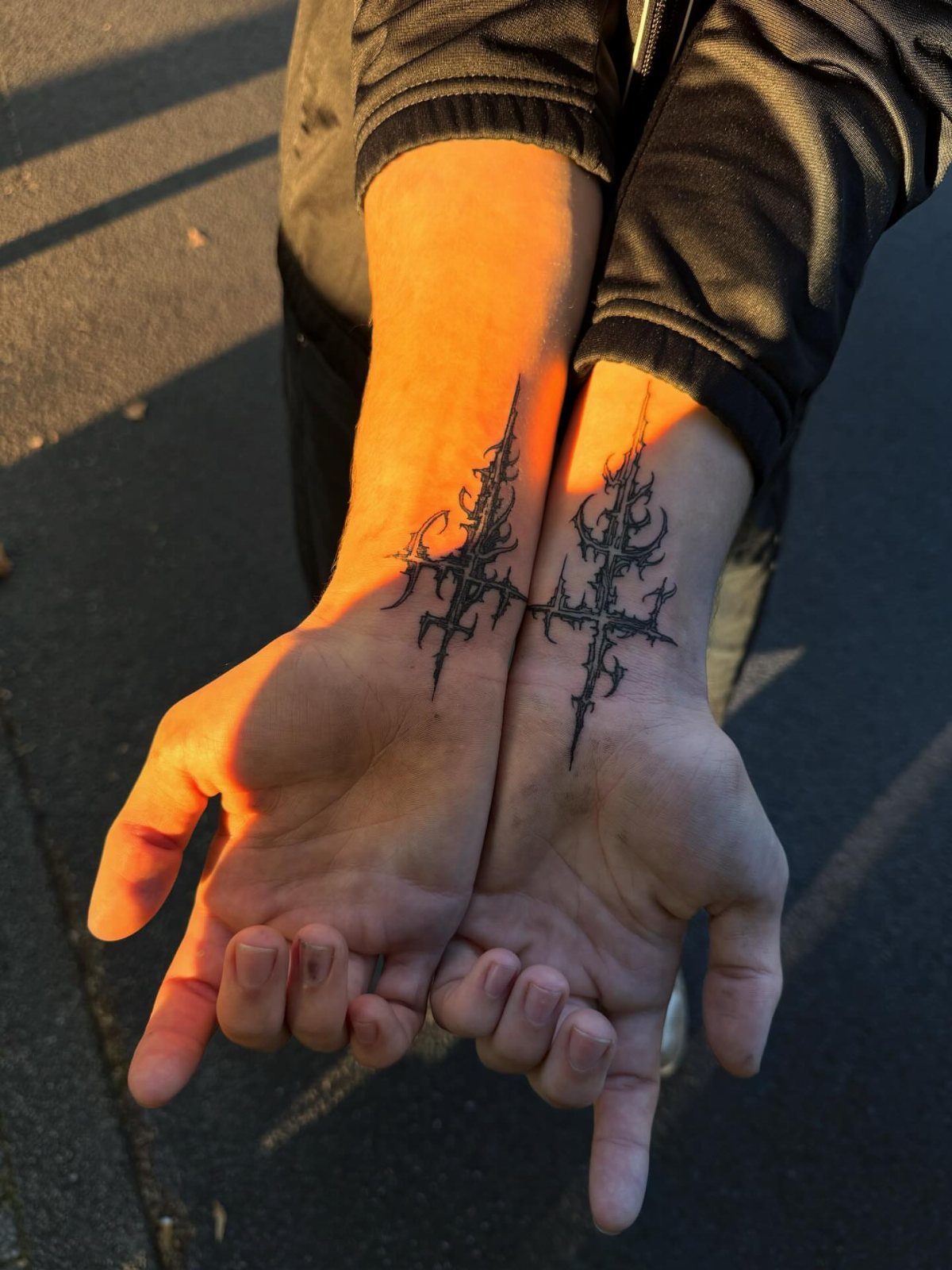 Two inner wrists featuring ornate black ink cross tattoos in golden sunlight.