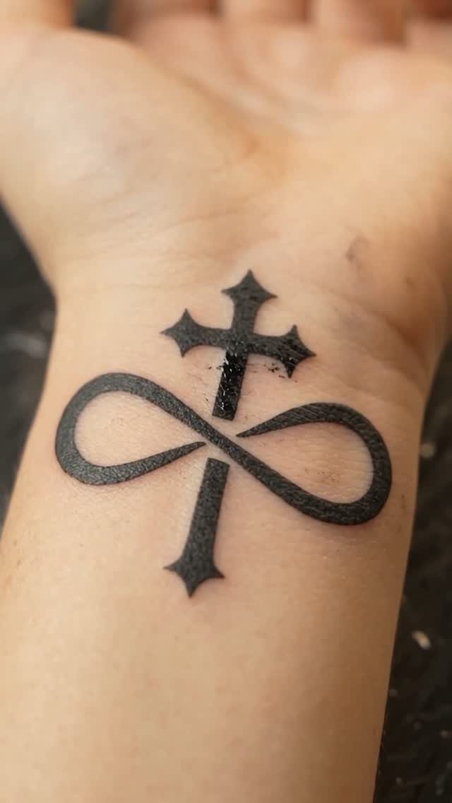 Black ink tattoo on a wrist combining a cross and an infinity symbol.
