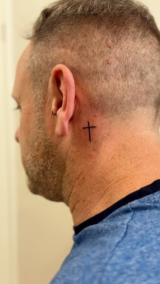 Small black cross tattoo on the side of a man's neck behind his ear