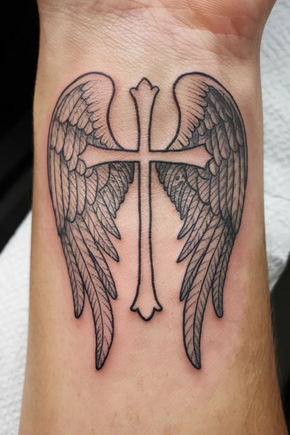 Cross tattoo with detailed angel wings on a person's inner wrist