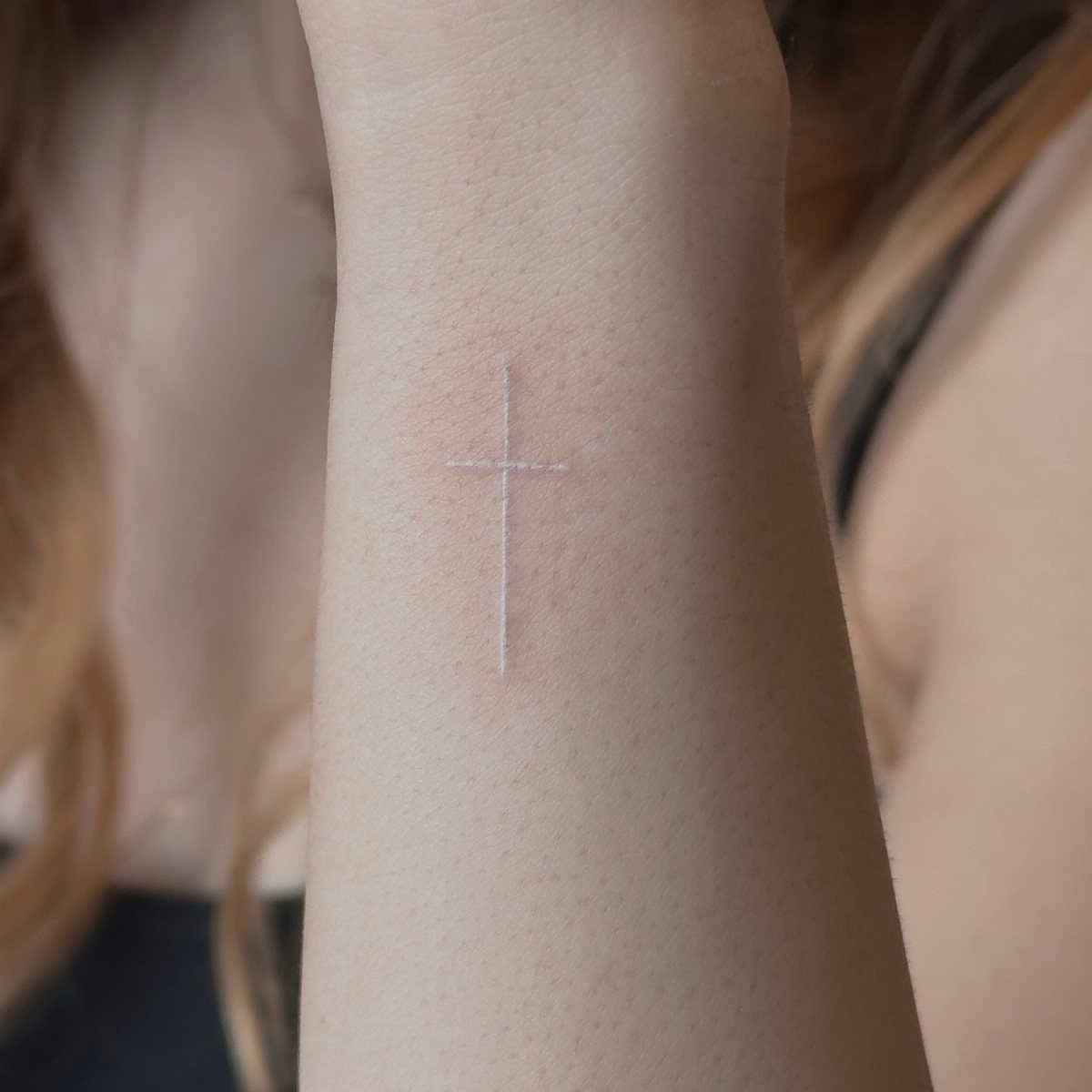 Small white ink cross tattoo centered on the inner wrist of a person.