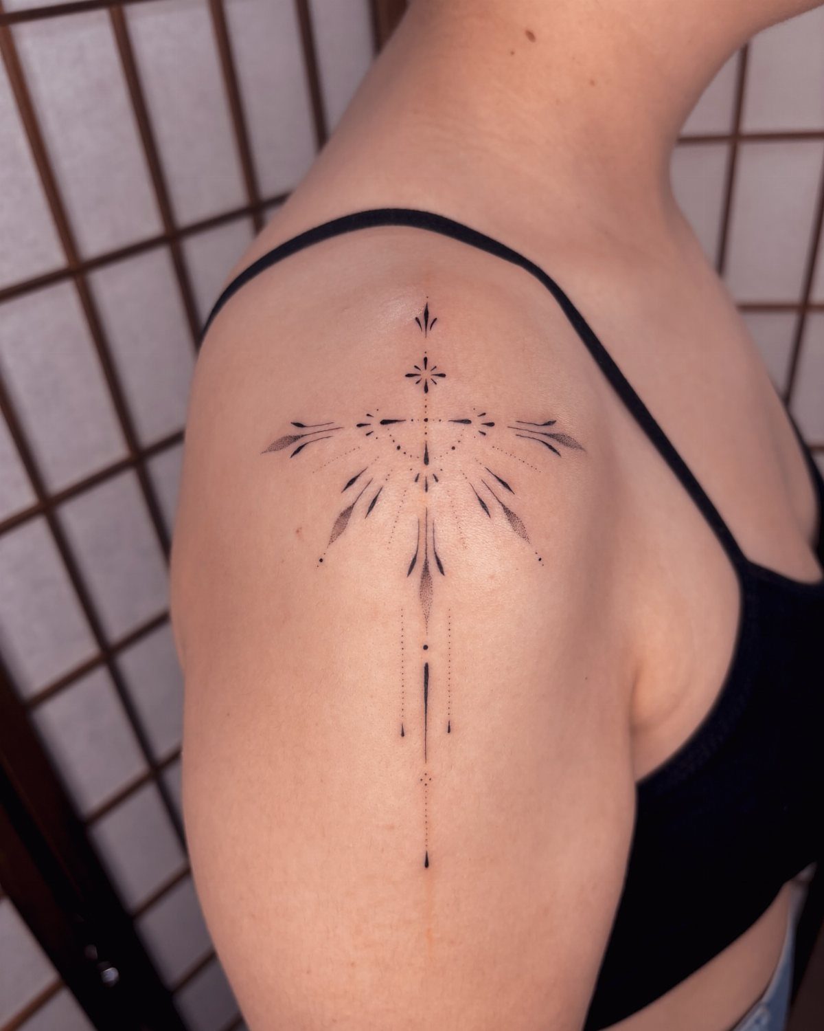 Geometric black ink cross tattoo with fine lines and dots on a shoulder