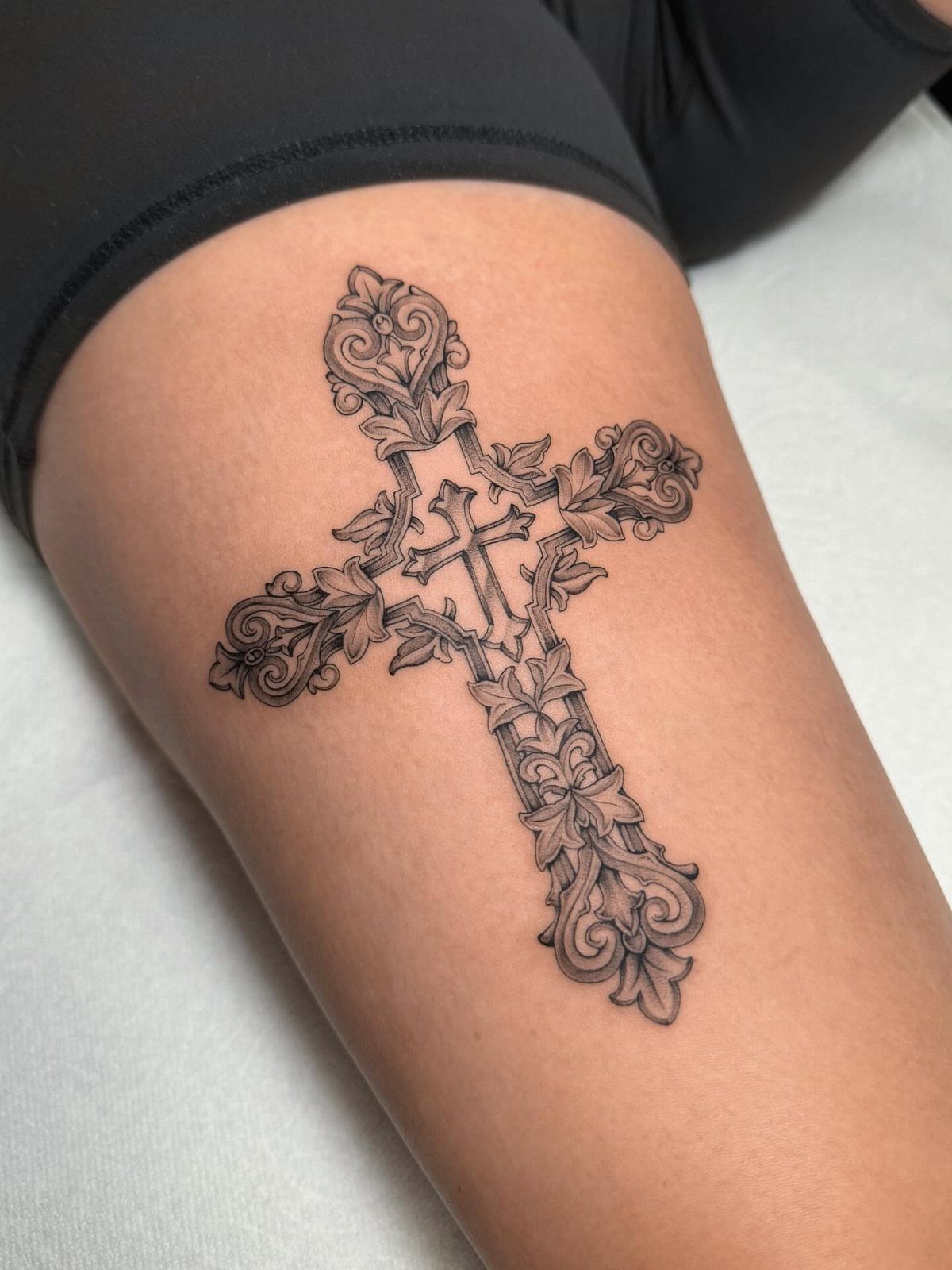 Ornate black ink cross tattoo with decorative scrolls on a person's thigh